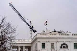 موقع بناء قاعة احتفالات جديدة أكبر من البيت الأبيض، يظهر رافعة تعمل على السطح، مع العلم الأمريكي يرفرف في الخلفية.