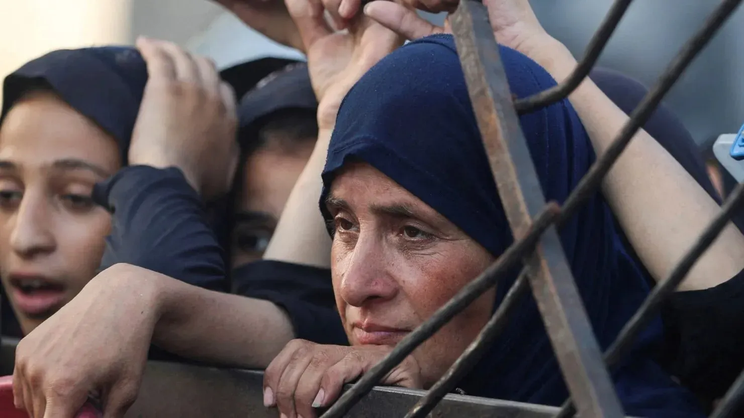 امرأة فلسطينية ترتدي حجابًا، تعبر عن مشاعر الحزن والقلق، محاطة بأشخاص آخرين، تعكس معاناتهم في ظل الأزمات الإنسانية في غزة.