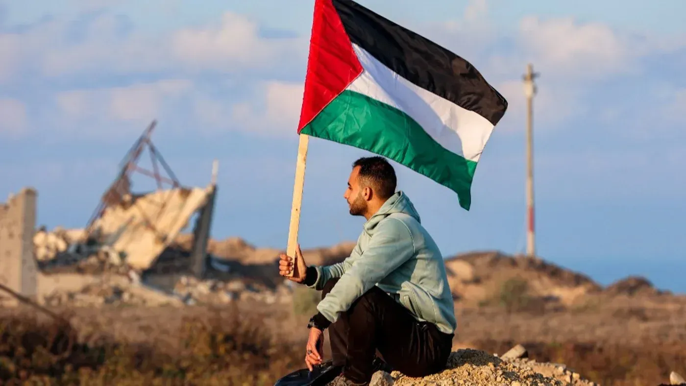 رجل يجلس على الأرض ممسكًا بعلم فلسطين، يطل على خلفية من الأنقاض، معبرة عن المعاناة في غزة.