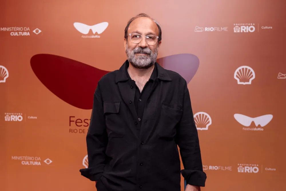 المخرج الإيراني أصغر فرهادي في مهرجان ريو السينمائي، مرتدياً نظارات وقميصاً أسود، مع خلفية تحمل شعار المهرجان وداعميه.