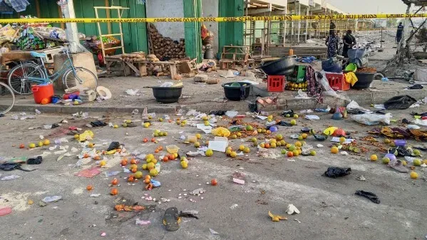 فوضى في سوق مزدحم في مايدوغوري بعد تفجيرات انتحارية، مع وجود حطام وفواكه متناثرة، بينما تواصل قوات الأمن جهودها في الموقع.