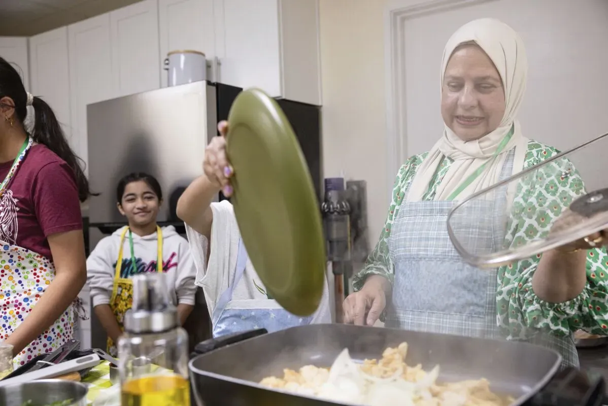 امرأة مسنّة ترتدي مئزرًا أزرق، تطهو أمام مجموعة من الأطفال في مخيم صيفي، حيث يتعلمون مهارات الطهي والحرف اليدوية.