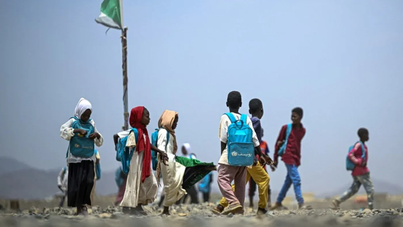 أطفال سودانيون يرتدون حقائب مدرسية، يسيرون في منطقة قاحلة، مع العلم السوداني في الخلفية، مما يعكس تأثير النزاع على التعليم.