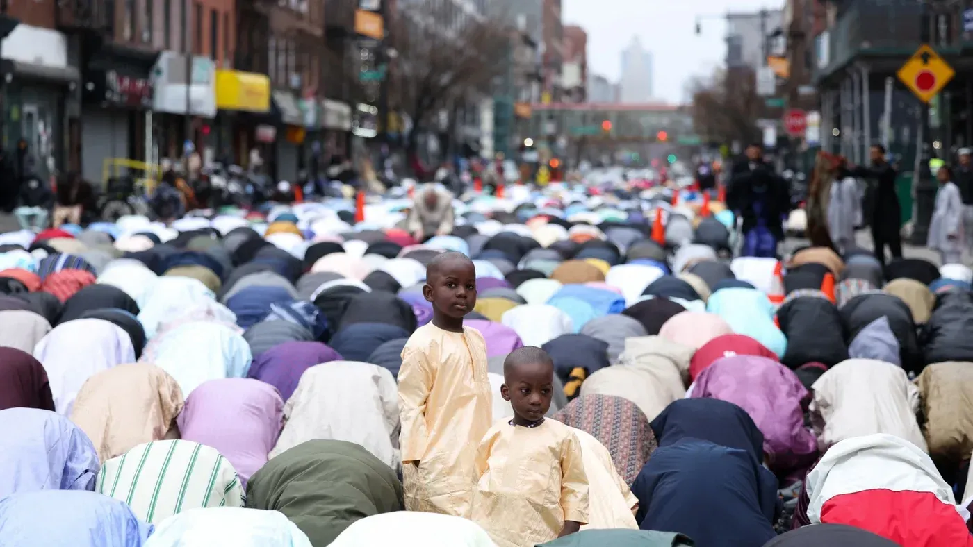 مجموعة من المسلمين يؤدون الصلاة في الشارع، مع وجود طفل صغير في المقدمة، تعكس مشهدًا من التعبير الديني في ظل تصاعد التوترات.