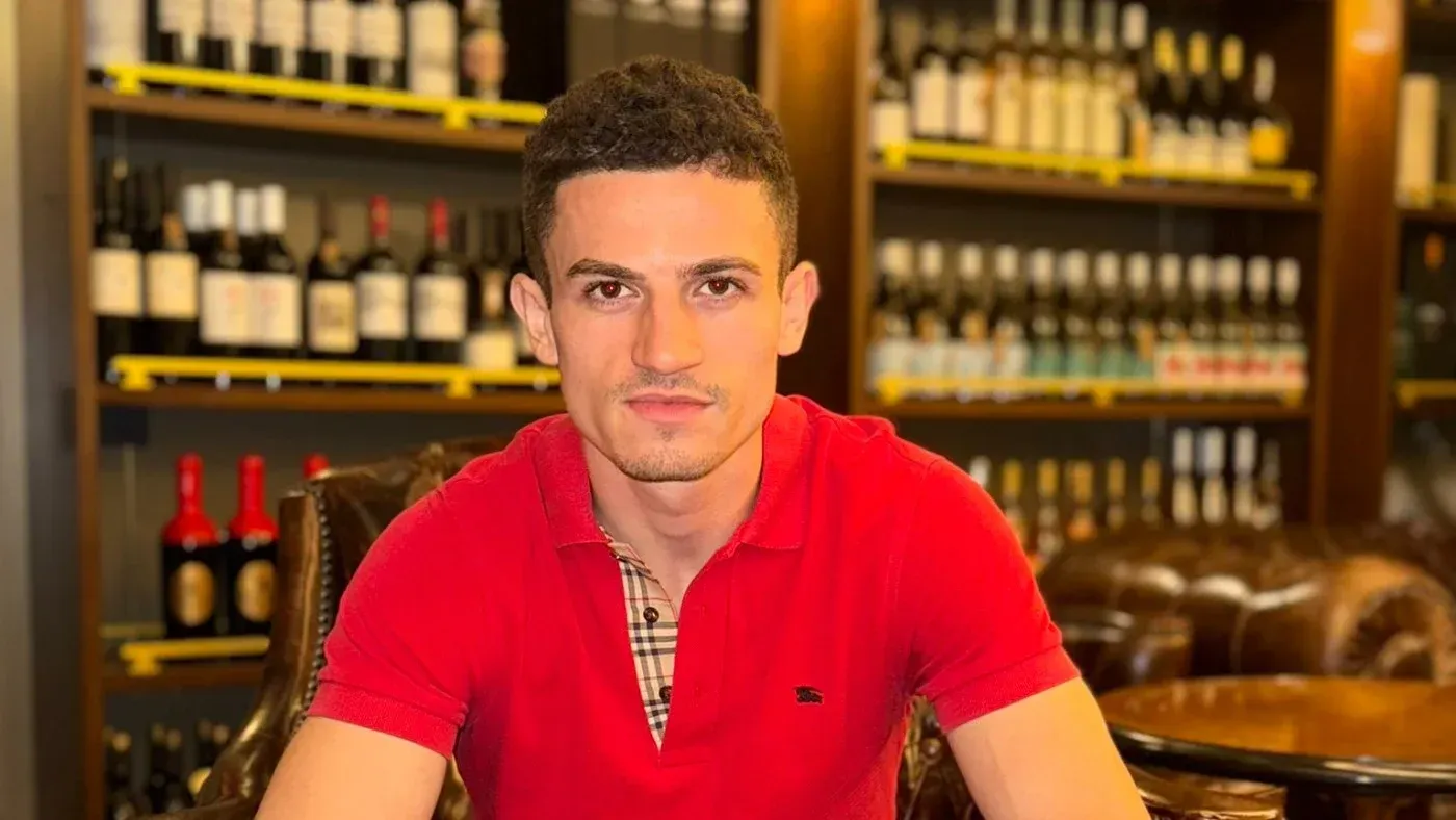 شاب يجلس في مطعم، يرتدي قميصًا أحمر، مع خلفية من زجاجات المشروبات، في سياق مناقشة حول إلغاء تأشيرة مؤثر يدعو لنشر الكراهية.