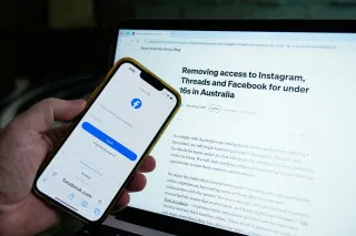شخص يحمل هاتفًا يعرض صفحة تسجيل الدخول إلى Instagram، وخلفه شاشة كمبيوتر تعرض مقالًا عن مضاعفة أستراليا الغرامات على منصات التواصل الاجتماعي لحظر حسابات الأطفال تحت 16 عامًا.