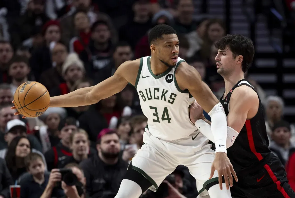 جيانيس أنتيتوكونمبو لاعب باكس يحمل الكرة ويواجه ضغط الدفاع من لاعب تريل بليزرز، مع جماهير متحمسة خلفهم في ملعب كرة السلة.