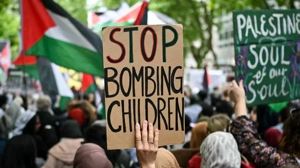 لافتة في مظاهرة تطالب بوقف قصف الأطفال، مع ظهور أعلام فلسطينية في الخلفية، تعكس مشاعر الاحتجاج ضد العنف في غزة.