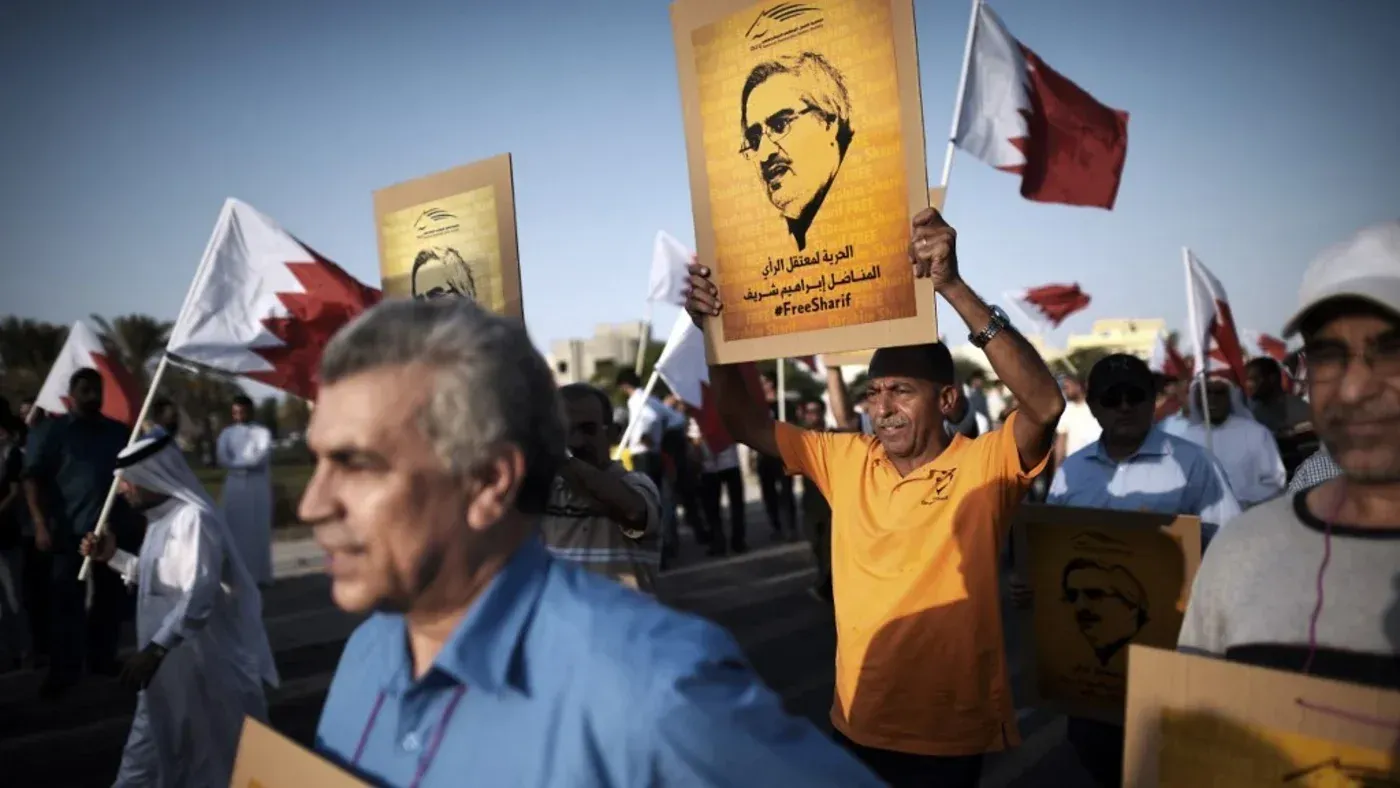 محتجون بحرينيون يحملون لافتات تطالب بالإفراج عن زعيم المعارضة إبراهيم شريف، وسط أعلام البحرين، تعبيراً عن دعمهم له.