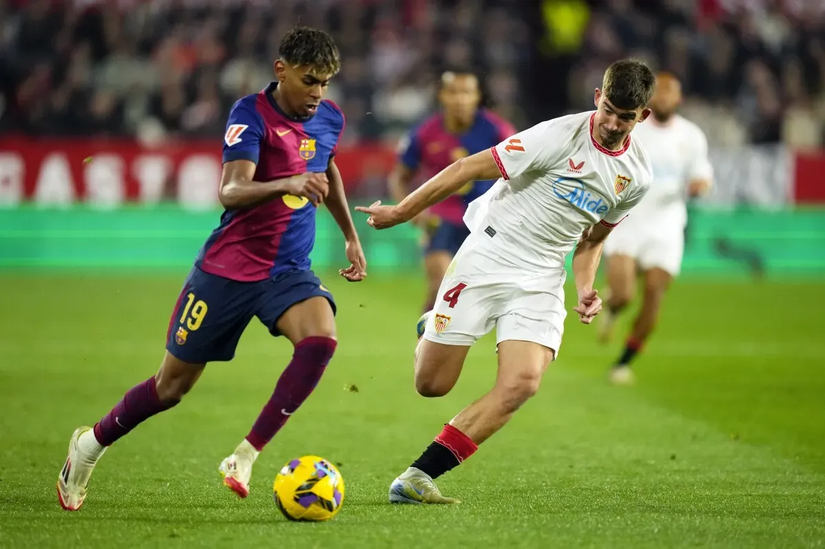لاعبا برشلونة وإشبيلية يتنافسان على الكرة في مباراة الدوري الإسباني، حيث يظهر لاعب برشلونة برقم 19 في قميص أزرق وأحمر.