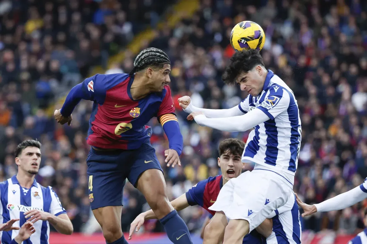 لاعبو برشلونة يتنافسون مع لاعبي ريال سوسيداد في مباراة كرة قدم، حيث يظهر لاعب برشلونة في المقدمة أثناء محاولة تسجيل هدف.