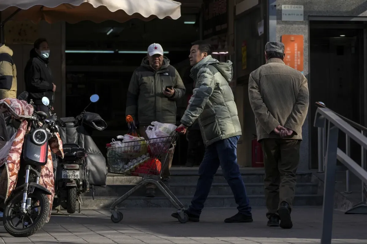 رجل يدفع عربة تسوق مليئة بالبقالة في حي سكني ببكين، مع وجود شخصين آخرين في الخلف، مما يعكس تأثير الانكماش الاقتصادي.