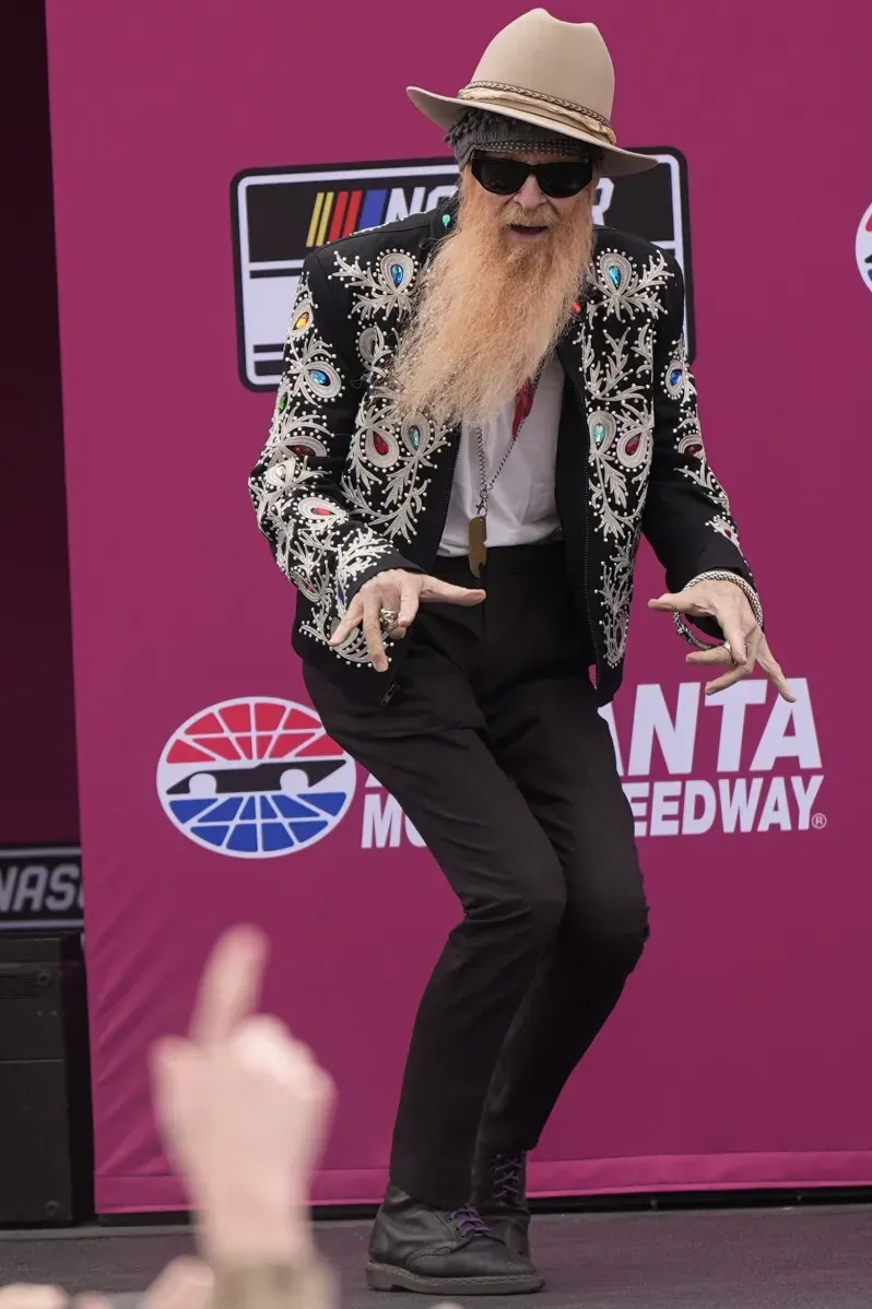 بيلي غيبونز، عازف الجيتار في فرقة ZZ Top، يؤدي بحماس مرتديًا بدلة مزخرفة وقبعة، خلفه خلفية وردية تحمل شعار ناسكار.