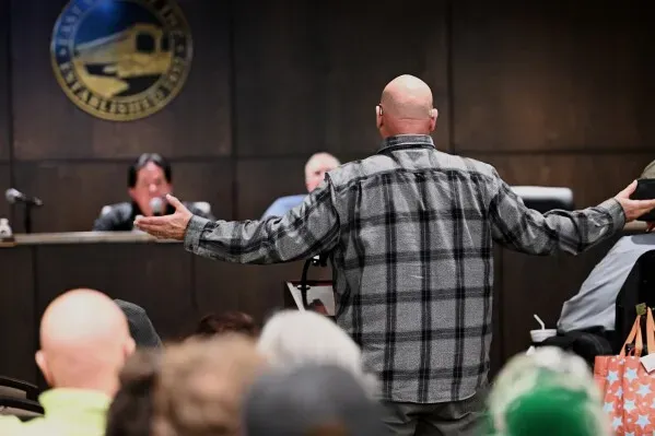 رجل يتحدث في اجتماع بلدي، مع جمهور غاضب خلفه، يعبر عن مخاوفه من إنشاء مراكز بيانات جديدة في مجتمعه.