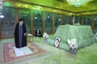 شخصية سياسية إيرانية تقف أمام ضريح مغطى بغطاء أخضر، مع صورة لشخصية معروفة وزهور بيضاء، في أجواء هادئة.