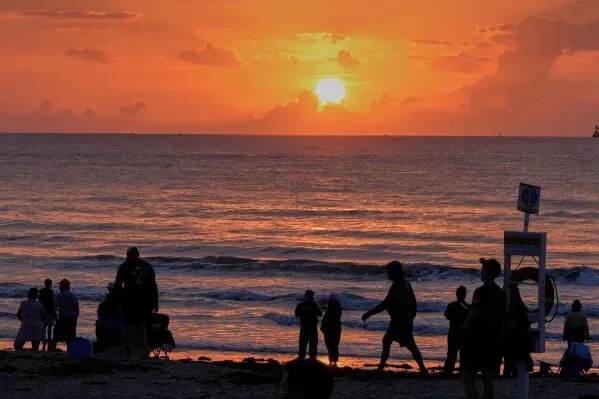 غروب الشمس يضيء السماء بلون برتقالي، مع silhouettes لأشخاص يستمتعون على الشاطئ، مما يعكس أجواء الاسترخاء بعد إطلاق صاروخ New Glenn.