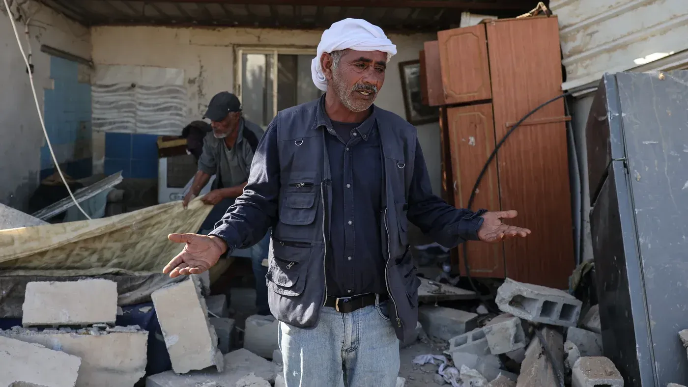 رجل فلسطيني يتحدث بقلق وسط أنقاض منزله المدمر، مع وجود أثاث مكسور وحطام حوله، معبرًا عن تأثير الاستيطان على حياته.