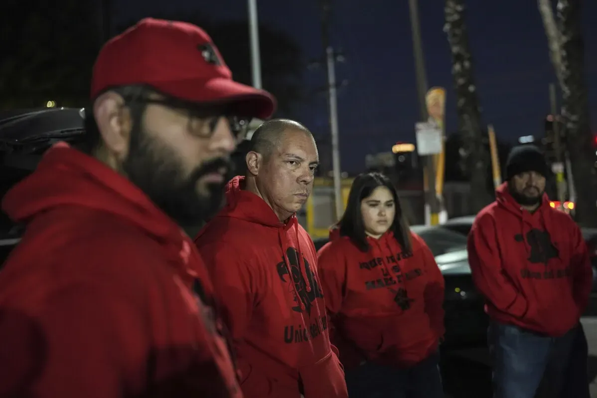 مجموعة من الأشخاص يرتدون سترات حمراء، يظهرون في اجتماع ليلي، مع تعبيرات جدية، يتحدثون عن قضايا الهجرة في بوسطن.