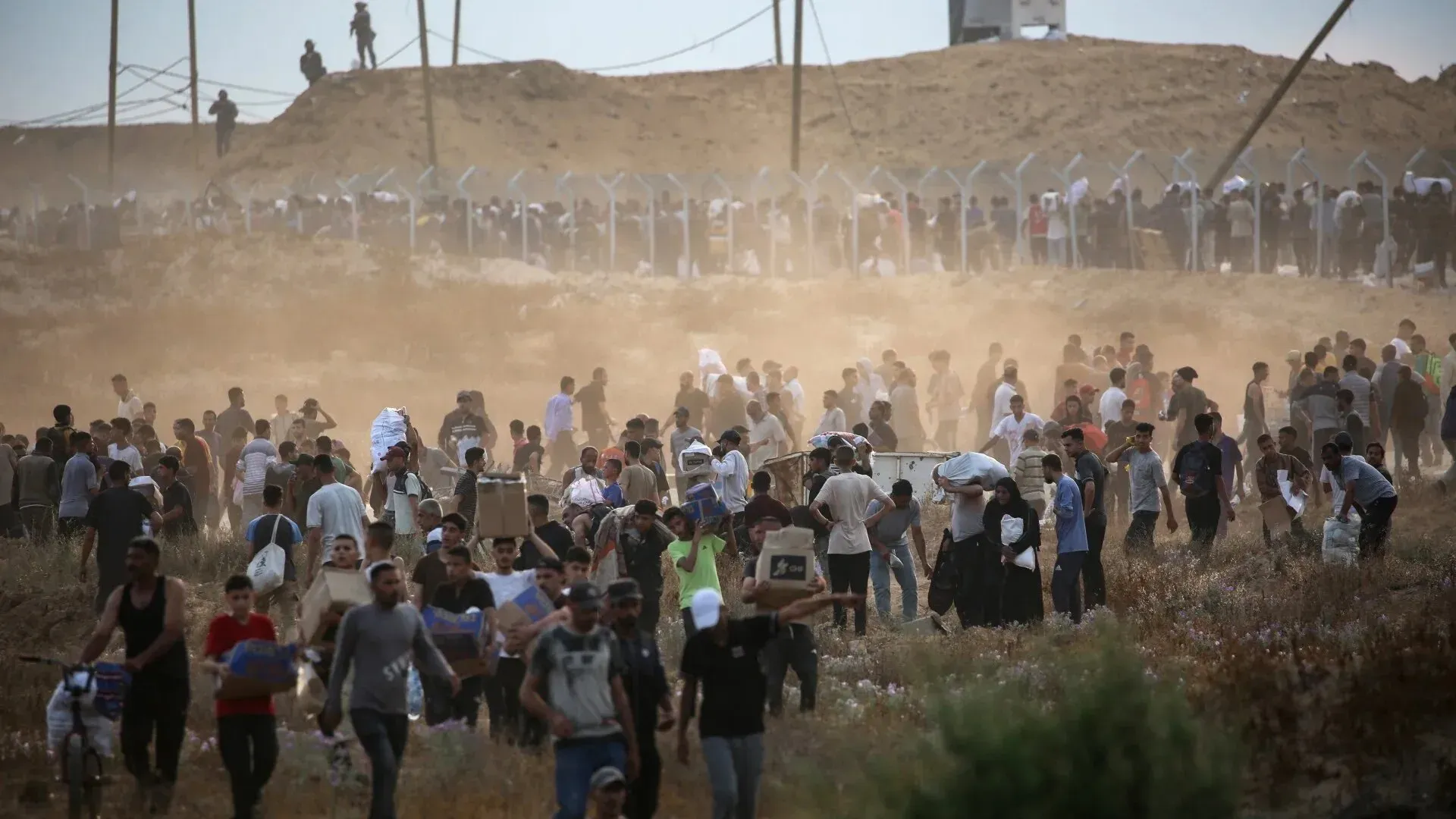 حشود من الفلسطينيين في غزة يحملون صناديق، يعبرون عبر منطقة ترابية، في ظل ظروف معيشية قاسية ونقص حاد في المواد الغذائية.