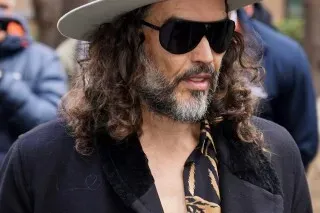 راسل براند، الممثل الكوميدي البريطاني، يرتدي نظارات شمسية وقبعة، ويظهر في محيط حضري، وسط قضايا قانونية جديدة تتعلق بالاعتداء الجنسي.