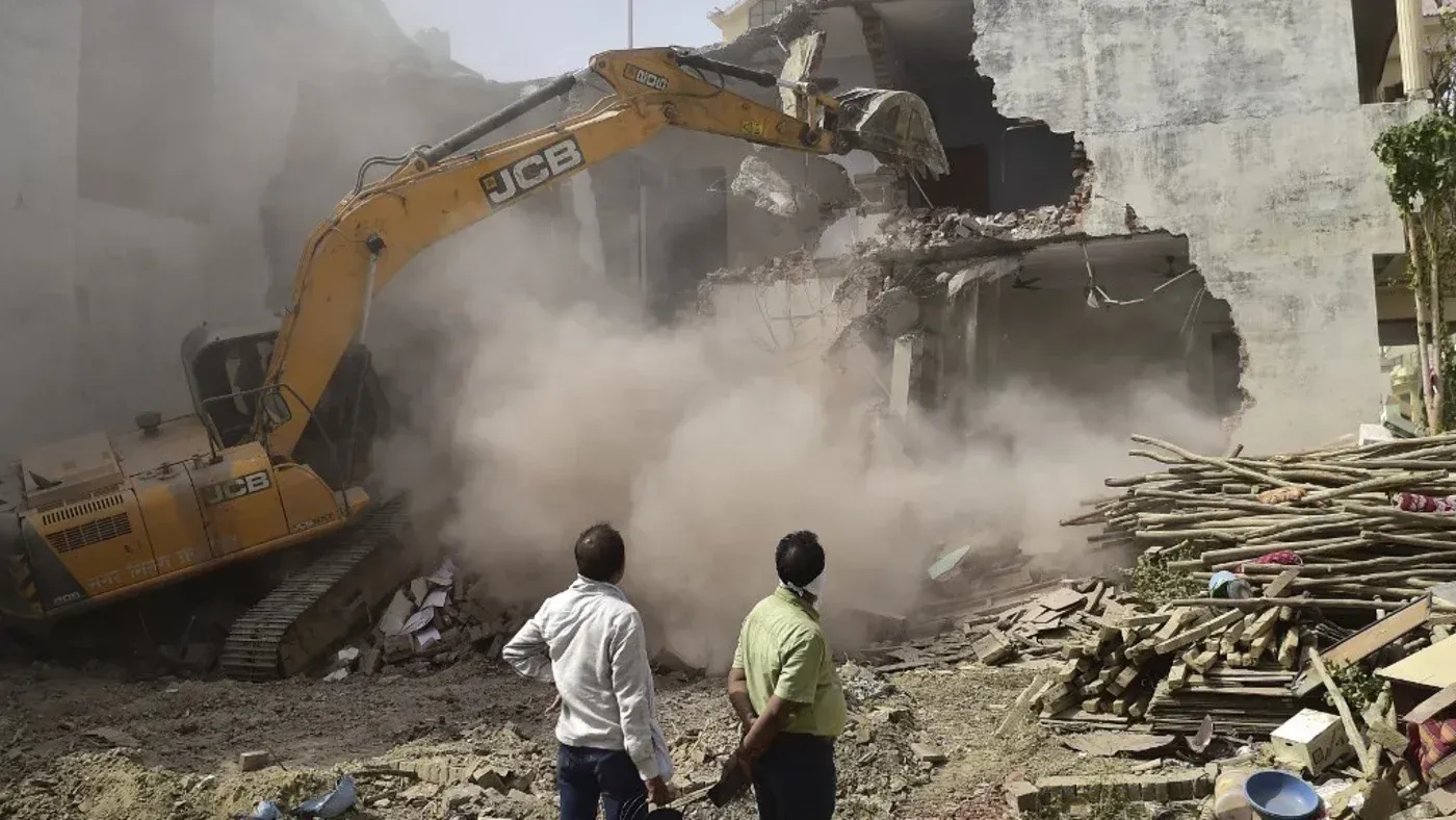 جرافة من طراز JCB تقوم بهدم مبنى، مما يثير الغبار ويظهر مشاهد توضح تأثير عمليات الهدم على المجتمعات المتضررة.