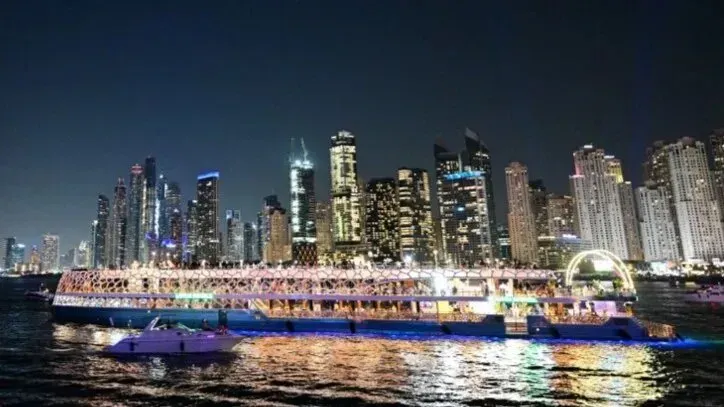 يظهر في الصورة قارب مضاء يجوب مياه دبي ليلاً، محاطاً بناطحات السحاب اللامعة، مما يعكس الحياة الليلية النابضة في المدينة.