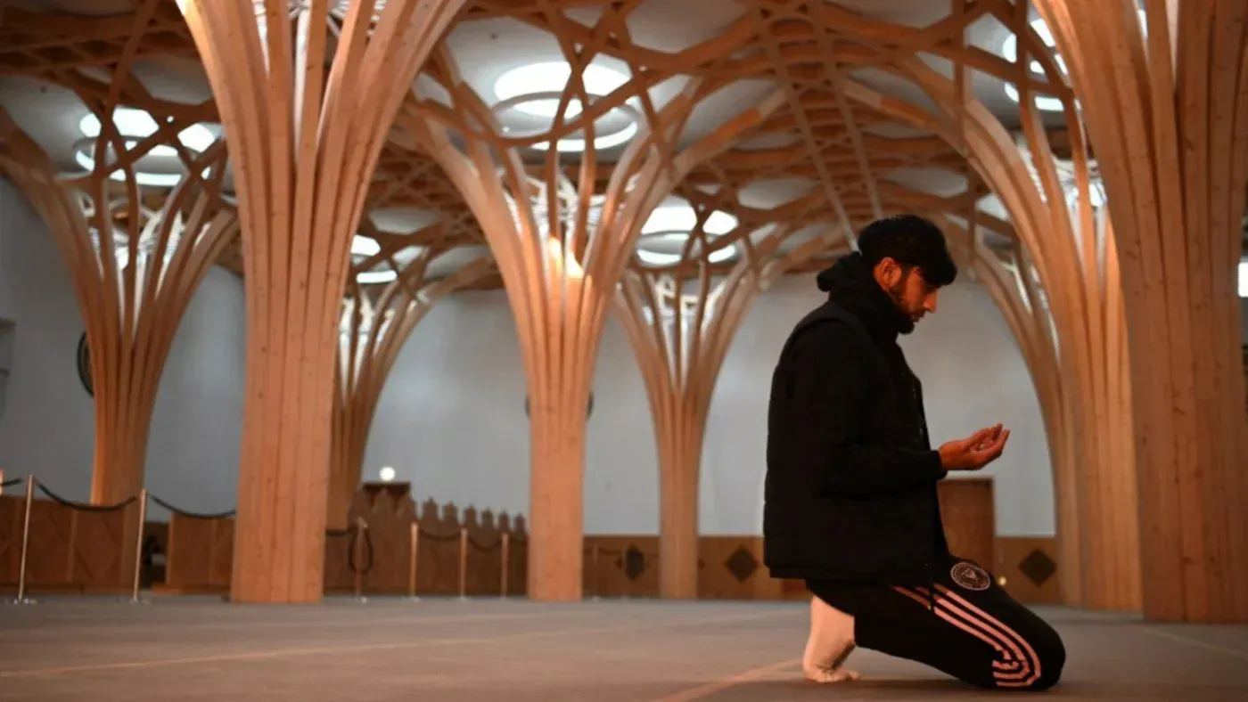 شاب مسلم يؤدي الصلاة في مسجد حديث، محاط بأعمدة خشبية معمارية، مما يعكس الاندماج الثقافي والديني في المجتمع البريطاني.