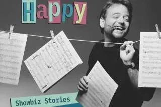 مارك شيمان مبتسم وهو يحمل أوراق موسيقية معلقة، مع عنوان "Happy" في الأعلى، يعكس مسيرته الفنية في عالم الترفيه.