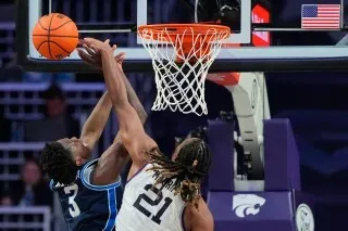 لاعب كرة سلة يحاول تسجيل نقطة تحت السلة بينما يتصدى له لاعب آخر، مع كرة سلة في الهواء، في مباراة مثيرة بين كانساس وبي واي يو.