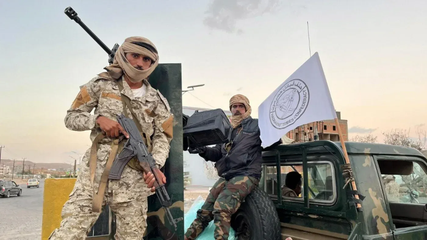 مقاتلان يرتديان زيًا عسكريًا، أحدهما يحمل سلاحًا والآخر يرفع علمًا، في سياق النزاع اليمني وتفكك الدولة.