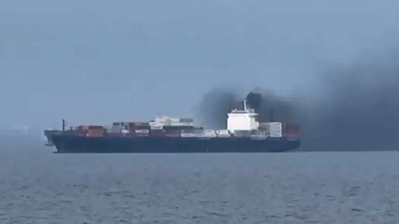 سفينة شحن كبيرة تبحر في البحر، حيث تتصاعد سحب من الدخان الأسود من محركاتها، مما يعكس التوترات في مضيق هرمز.