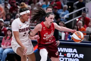 لاعبة كرة السلة كيتلين كلارك من Indiana Fever تتقدم بالكرة خلال مباراة في دوري WNBA وسط منافسة قوية، مع تسليط الضوء على تحديات الكراهية الرقمية التي تواجه اللاعبات.