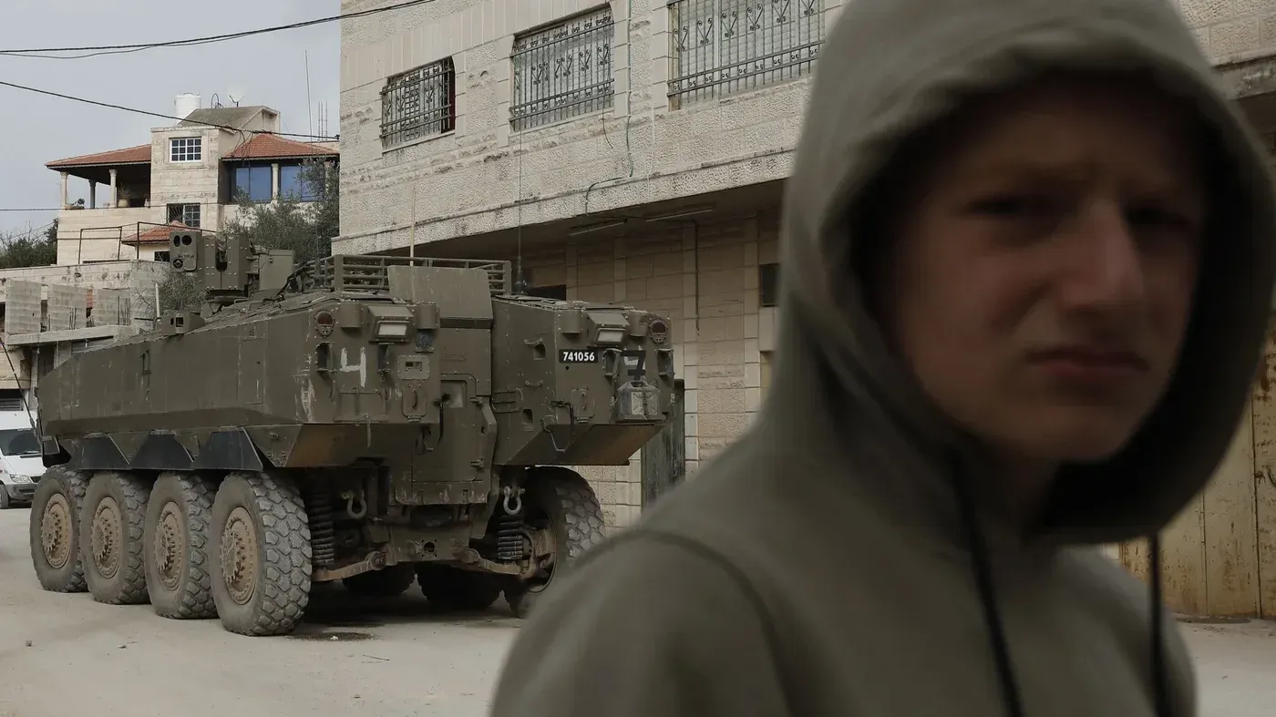 طفل يرتدي سترة رمادية يقف في الشارع بالقرب من مركبة عسكرية إسرائيلية، مما يعكس التوترات الحالية في المنطقة.