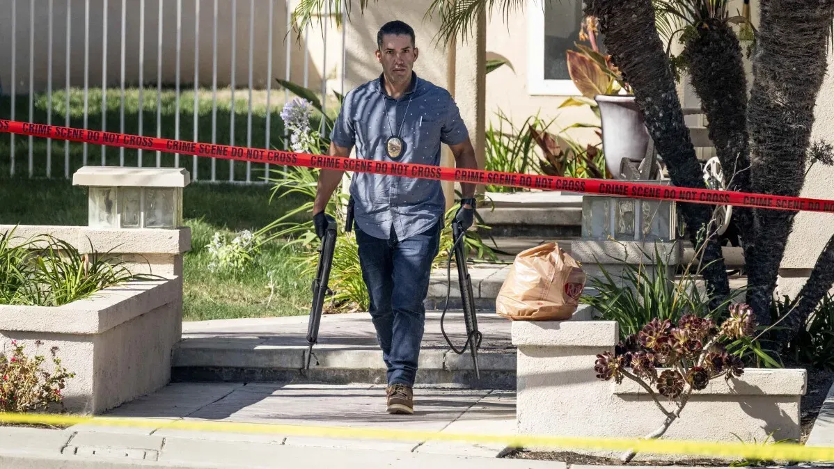 شرطي يحمل بندقية ويخرج من موقع جريمة محاط بشريط تحذيري، مما يشير إلى حادث إطلاق نار في سانتا آنا، كاليفورنيا.