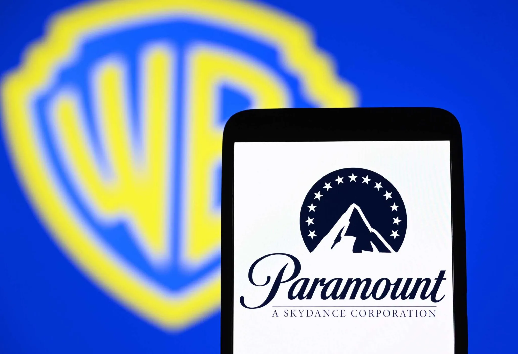 شعار Paramount على شاشة هاتف ذكي أمام شعار Warner Bros. Discovery، يعكس صفقة اندماج ضخمة في صناعة الترفيه الأمريكية وتأثيرها على السوق.