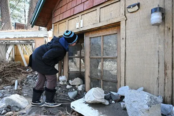 امرأة ترتدي معطفاً تنظر من نافذة منزل محاط بالحطام والصخور بعد الفيضانات في رايتوود، كاليفورنيا.