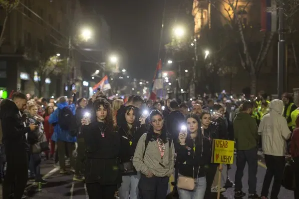 محتجون في بلغراد يحملون هواتف مضاءة خلال مسيرة ضد الفساد، مع لافتات تطالب بالتحقيق في استخدام سلاح صوتي محظور.