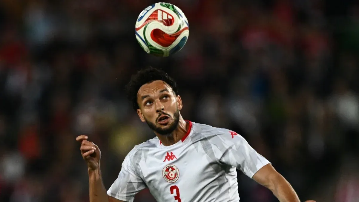 لاعب كرة قدم تونسي يرتدي قميص المنتخب الوطني، يرفع رأسه ليلتقط كرة متجهة نحو الأعلى، وسط أجواء حماسية في الملعب.