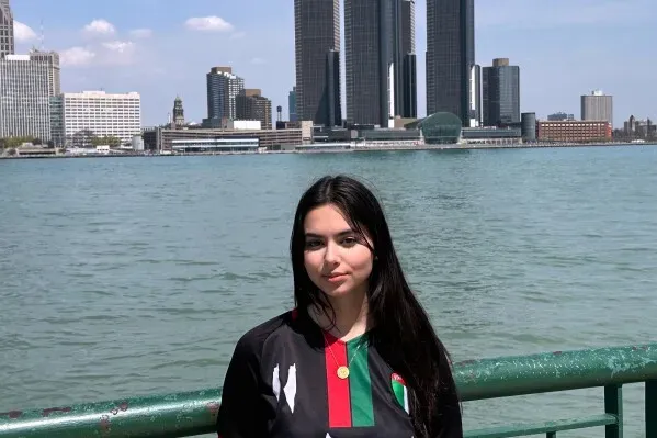 امرأة شابة ترتدي قميصًا رياضيًا، تقف أمام نهر مع منظر لمدينة ديترويت، حيث تظهر المباني الشاهقة في الخلفية.