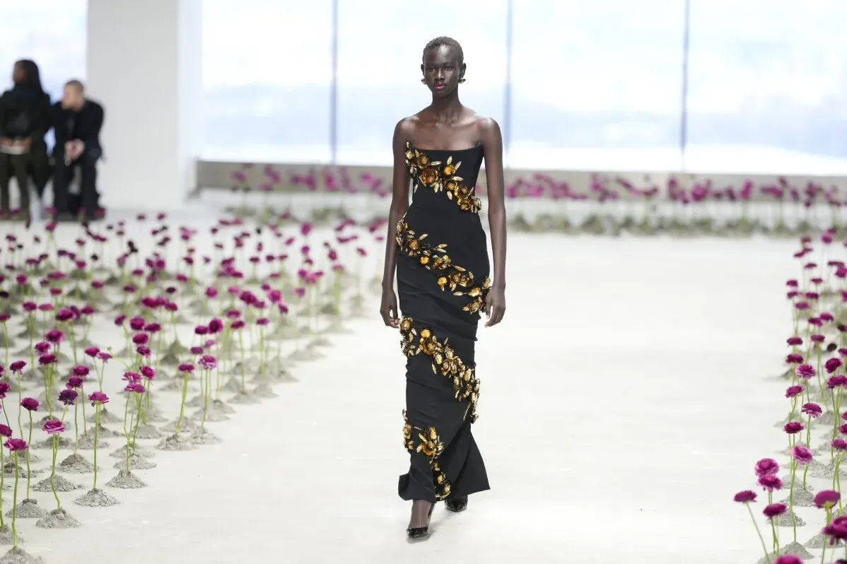 عارضة أزياء ترتدي فستانًا أسود مزينًا بأزهار ذهبية، تسير على مدرج محاط بزهور رانونكولوس بلون النبيذ في عرض كارولينا هيريرا.