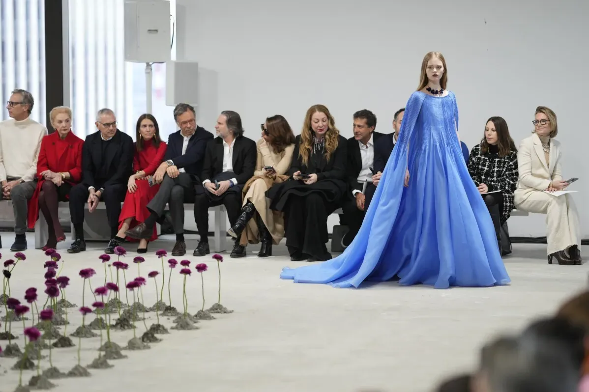 عارضة أزياء ترتدي فستانًا أزرقًا طويلًا وتظهر على المدرج في عرض أزياء كارولينا هيريرا، مع جمهور من الشخصيات البارزة في الخلفية.