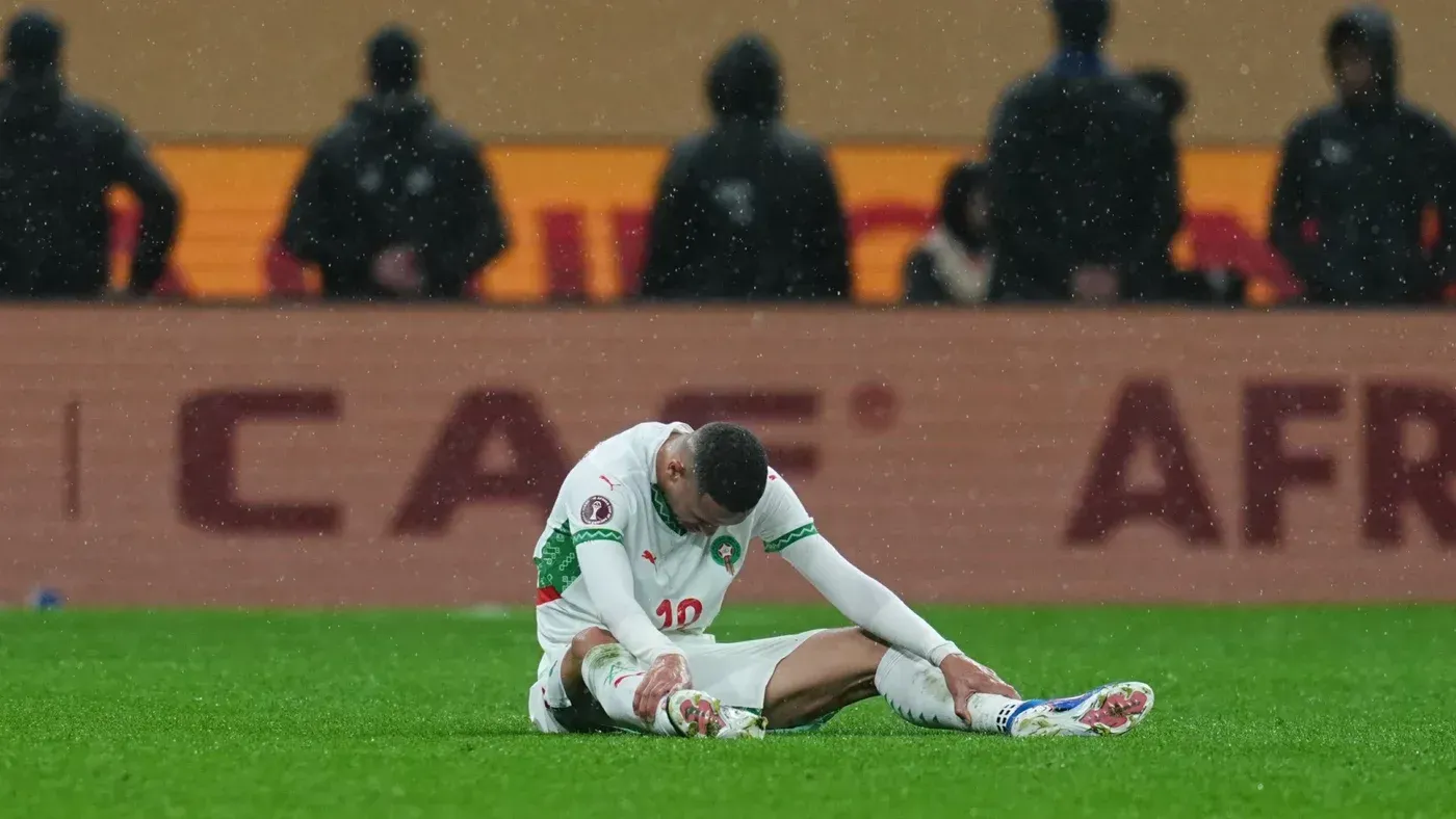 لاعب مغربي يجلس على الأرض في حالة إحباط بعد هزيمة فريقه في نهائي كأس الأمم الإفريقية، مع خلفية تشير إلى الحدث الرياضي.