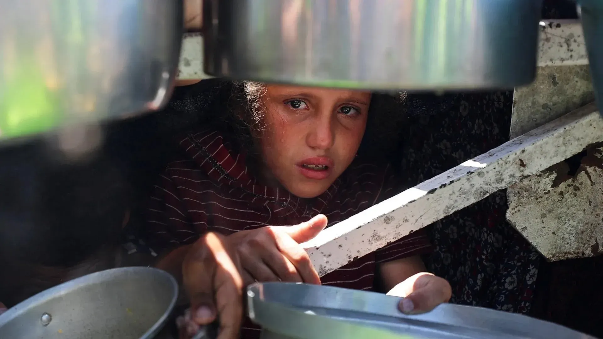 طفل فلسطيني يظهر بملامح حزينة وهو يجلس تحت مجموعة من الأواني، يعكس معاناة الجوع وسوء التغذية في غزة.