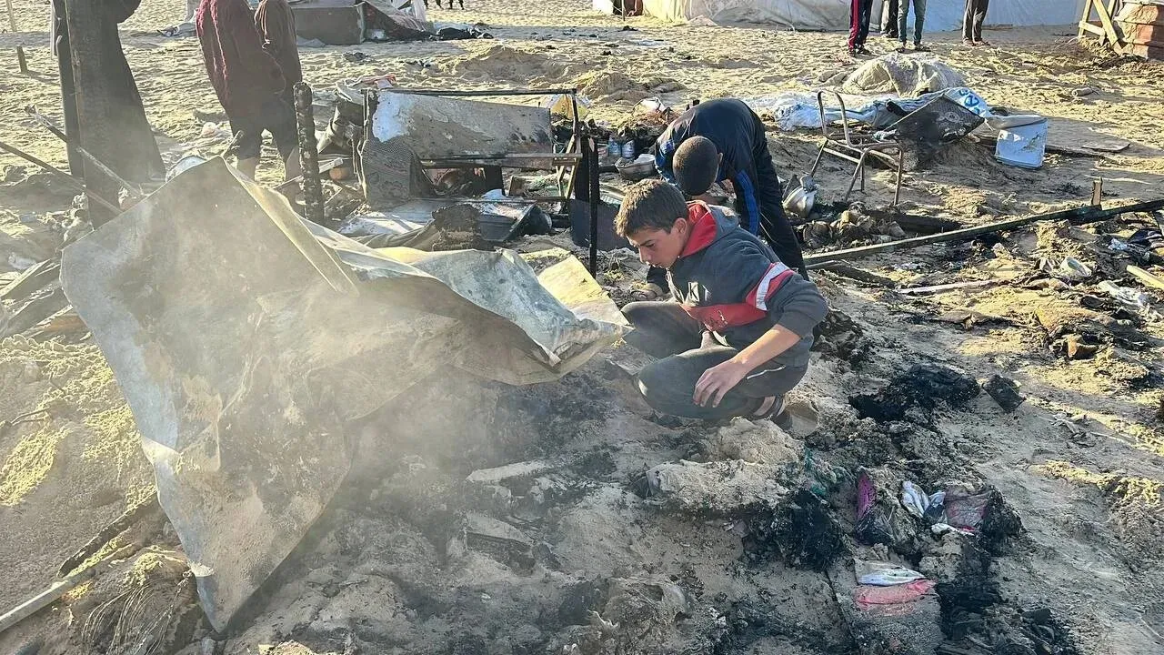 مشهد مروع يظهر شابًا يبحث بين الأنقاض المتفحمة في منطقة المواصي بخان يونس بعد الغارات الإسرائيلية، مع وجود خيام محترقة في الخلفية.