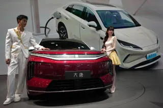 عرض لشخصين يقفان بجانب سيارة كهربائية حمراء من شركة BYD، مع سيارة بيضاء في الخلفية، في سياق حديث حول العلاقات الاقتصادية الصينية الأمريكية.