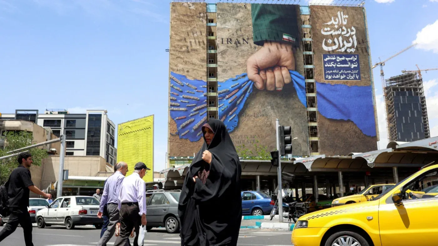لوحة إعلانات ضخمة في طهران تظهر يدًا ممسكةً بعلم إيران، مع خلفية زرقاء وأسلحة، بينما يمر المارة والسيارات في الشارع.