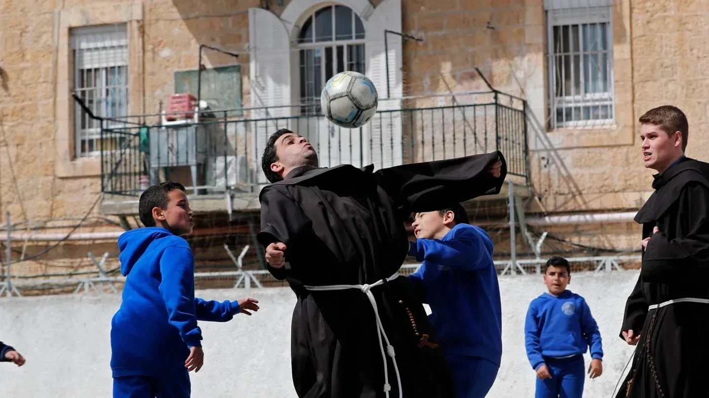 طلاب ومعلمون في مدرسة مسيحية في القدس يلعبون كرة القدم في ساحة المدرسة، تعبيرًا عن روح التعاون والمرح وسط التحديات التعليمية.