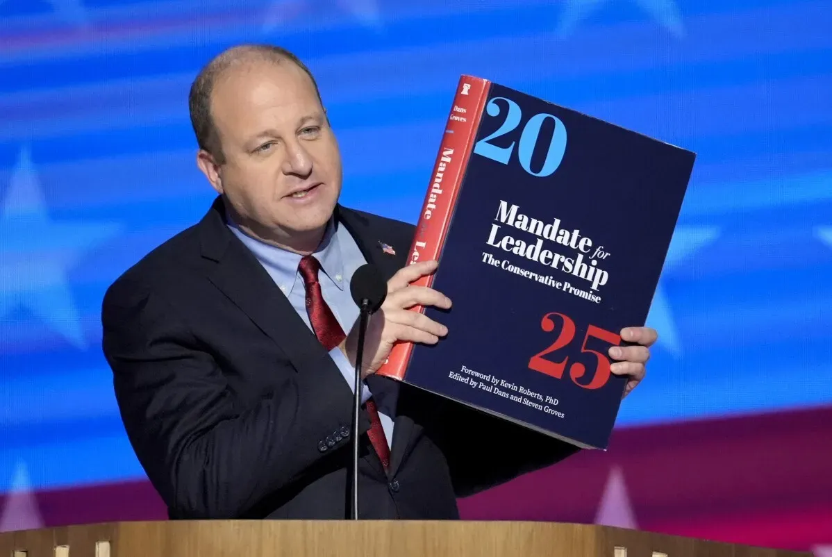 جاريد بوليس، حاكم كولورادو، يحمل كتاب \"Mandate for Leadership\" خلال مؤتمر صحفي، مشيرًا إلى إلغاء التنظيمات غير الضرورية.