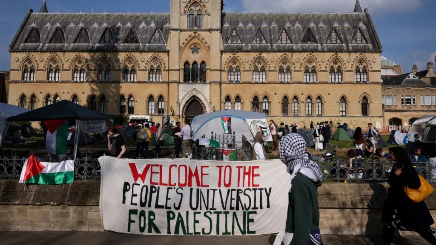 تظاهرة أمام جامعة بريطانية تحمل لافتة "مرحبًا بكم في جامعة الشعب لفلسطين"، مع وجود خيام وأعلام فلسطينية، تعبيرًا عن التضامن.