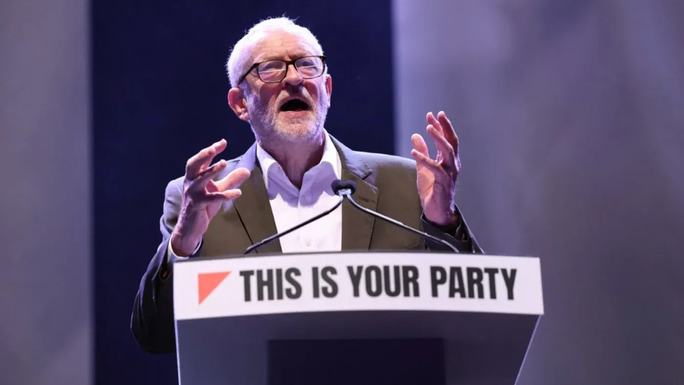 جيريمي كوربين يتحدث في مؤتمر، معبرًا عن انتقاده لحكومة حزب العمال بشأن عقد مع شركة بالانتير، مع خلفية تحمل شعار "هذه هي حزبك".